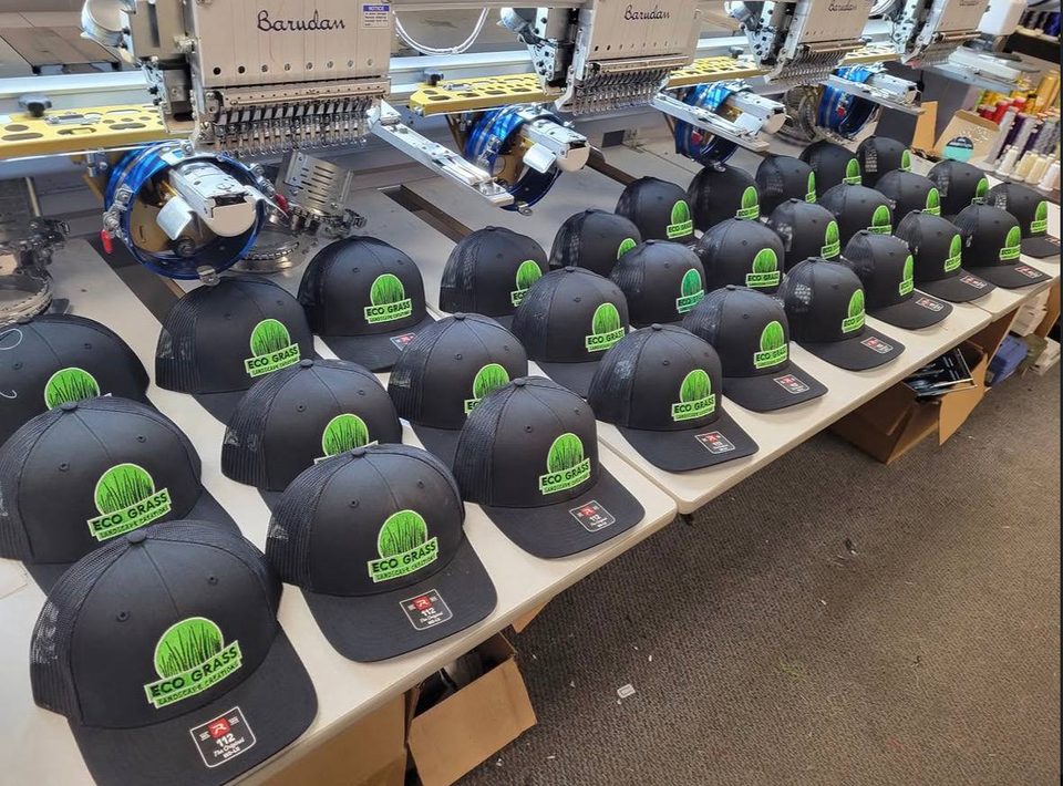 Rows of black trucker hats with a green landscaping logo on the embroidery machine table